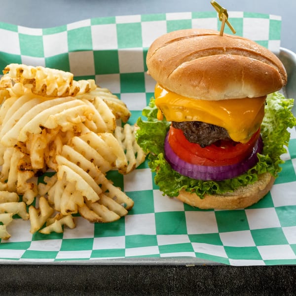 A cheeseburger with lettuce, tomato, and onion, served with waffle fries on checkered paper.