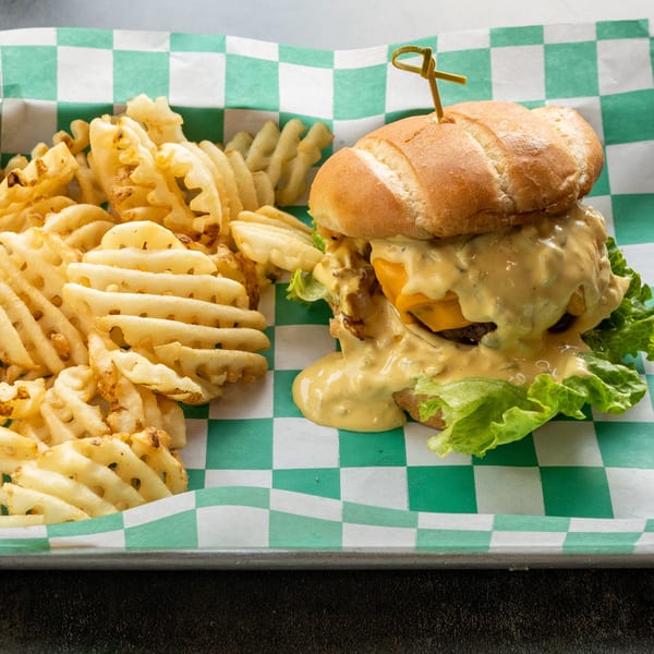 A burger with melted cheese and sauce, lettuce, and waffle fries on a green and white checkered paper.